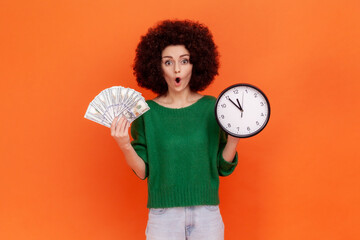 Shocked astonished woman with Afro hairstyle wearing green casual style sweater holding wall clock...