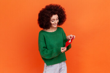 Portrait of positive female woman with Afro hairstyle wearing green casual style sweater standing holding red envelope, reading romantic letter. Indoor studio shot isolated on orange background.