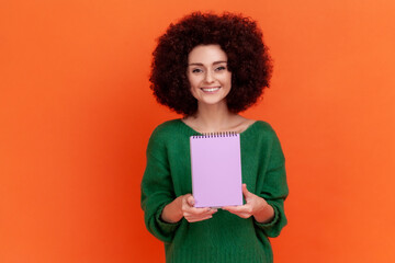 Portrait of satisfied woman with Afro hairstyle in green casual style sweater standing showing paper notebook, looking at camera, happy expression. Indoor studio shot isolated on orange background.