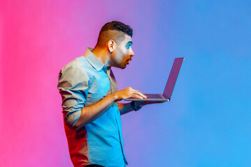 Side view of man in shirt doing freelance job on laptop, typing email or surfing internet, looking...