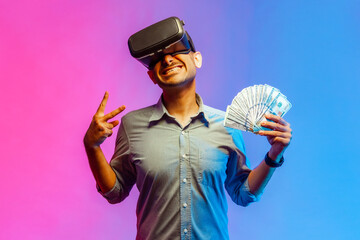 Portrait of smiling man in shirt holding dollars, illusion of rich millionaire, playing virtual reality game, showing v sign. Indoor studio shot isolated on colorful neon light background. - obrazy, fototapety, plakaty