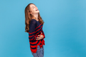 Hysterical laughter. Happy young woman wearing striped casual style sweater laughing out loud and holding belly, amused by humorous joke. Indoor studio shot isolated on blue background. - obrazy, fototapety, plakaty