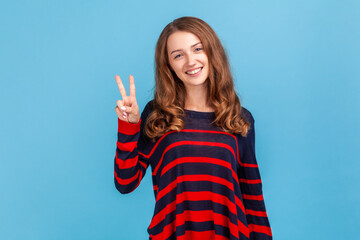 Happy satisfied woman wearing striped sweater showing v sign symbol of peace with fingers, looking at camera with toothy smile, celebrating victory. Indoor studio shot isolated on blue background.