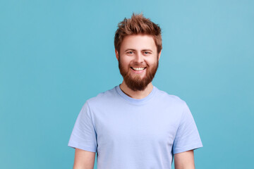 Obraz premium Portrait of bearded man looking at camera with toothy smile and happy facial expression, being in good mood, rejoicing great results. Indoor studio shot isolated on blue background.