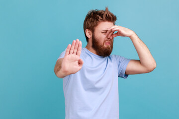 Portrait of handsome bearded man standing, pinching nose and rejecting or banning, showing stop ban gesture with palm, disgusting expression. Indoor studio shot isolated on blue background.