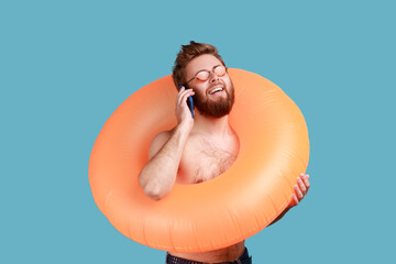 Portrait of overjoyed young adult bearded man standing with orange rubber ring and talking phone,...