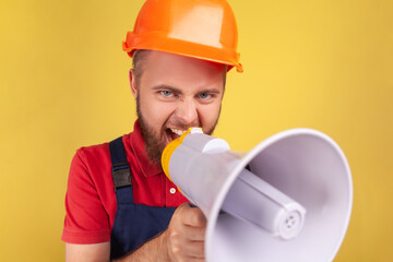 Angry aggressive bearded worker wearing protective helmet and blue overalls holding megaphone and screaming, point of view photo. Indoor studio shot isolated on yellow background.