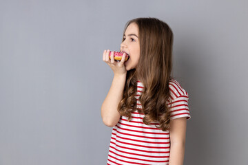 Portrait of hungry charming little girl wearing striped T-shirt standing biting doughnut and...