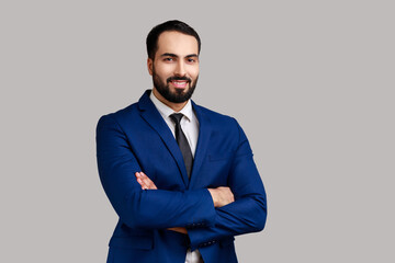 Optimistic young adult bearded businessman standing with crossed arms and looking at camera with...