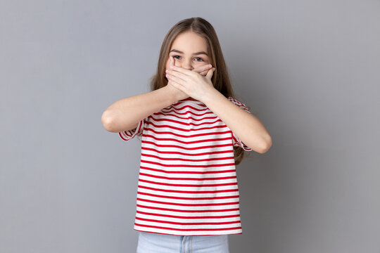 Portrait of dark haired little girl wearing striped T-shirt put hands on mouth, looking with fear in her eyes, keeping terrible secret. Indoor studio shot isolated on gray background.