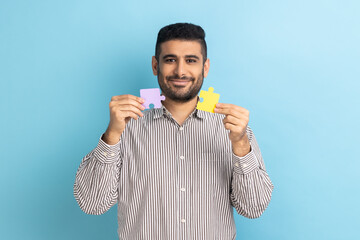 Portrait of optimistic bearded businessman holding yellow and purple puzzle pieces, solving tasks,...