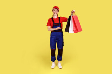 Beautiful optimistic courier woman standing with shopping bags in hands, showing presenting parcels, expressing happiness, wearing overalls and cap. Indoor studio shot isolated on yellow background.