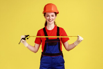 Portrait of woman builder holding roulette for measuring, looking at camera with happy facial expression, wearing overalls and protective helmet. Indoor studio shot isolated on yellow background.