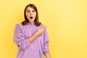Portrait of excited beautiful young adult woman standing and pointing at copyspace with surprised face, wearing purple hoodie. Indoor studio shot isolated on yellow background.