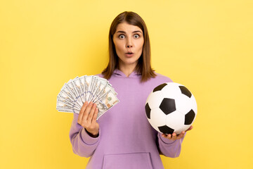 Portrait of woman with open mouth holding soccer ball and fan of dollars, amazed of football...