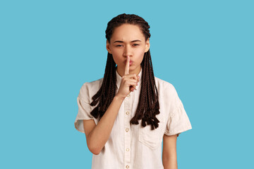 Woman with black dreadlocks shushes with angry face, makes taboo gesture, tells to be quiet, demands not to spread rumors, scolds person being too loud. Indoor studio shot isolated on blue background.