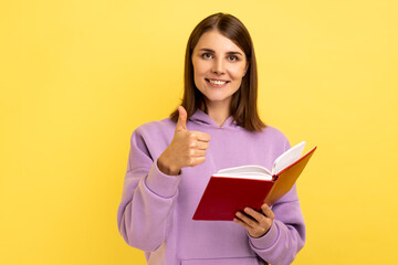 Portrait of happy satisfied woman showing thumbs up gesture holding and reading book, likes genre and interesting plot, wearing purple hoodie. Indoor studio shot isolated on yellow background.