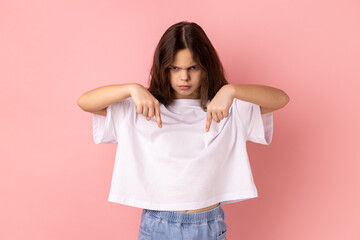 Portrait of strict serious little girl wearing white T-shirt pointing down, gesturing below with...
