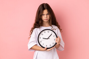 Portrait of little girl wearing white T-shirt holding wall clock, being unhappy, deadline, being sad, not finished her task in time. Indoor studio shot isolated on pink background.