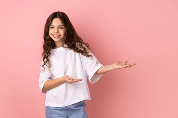 Portrait of kind-hearted generous little girl wearing white T-shirt showing welcome gesture and...