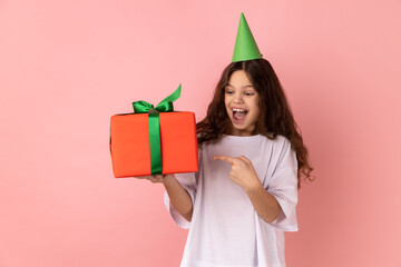 Look at my present. Little girl wearing white T-shirt and party cone on head and pointing at gift box, smiling to camera, boasting birthday surprise. Indoor studio shot isolated on pink background.