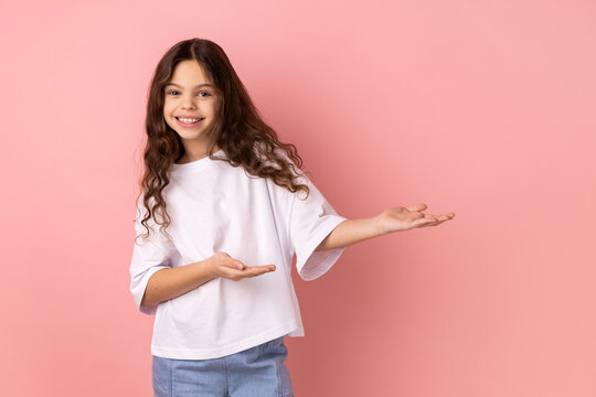 Portrait of kind-hearted generous little girl wearing white T-shirt showing welcome gesture and empty space on wall for your best advertising. Indoor studio shot isolated on pink background.