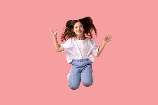 Portrait of happy delighted positive little girl wearing white T-shirt jumping in air with raised arms, feeling energetic and lively. Indoor studio shot isolated on pink background.