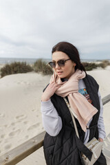Stylish beautiful woman with vintage sunglasses in fashionable clothes with a scarf, vest, and thick jacket walks on the beach on a wooden walkway by the ocean