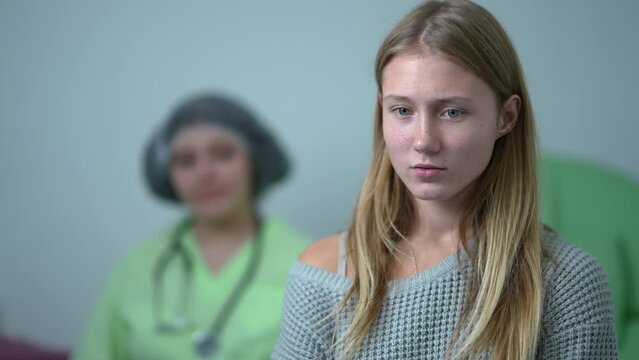 Unsure anxious teenage girl standing on the right thinking with blurred doctor waiting at background. Portrait of scared stressed Caucasian teenager on appointment with gynecologist in hospital