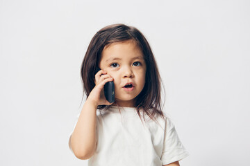 a little girl of preschool age, sitting on a white background in a white T-shirt, enthusiastically talking on the phone, turning half-side to the camera. The topic of the conversation on the phone