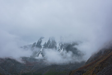 Dark atmospheric landscape with high snow mountains in dense fog in rainy weather. Large snowy mountain range in thick fog in dramatic overcast. Snow-covered rocky mountains in low clouds during rain.
