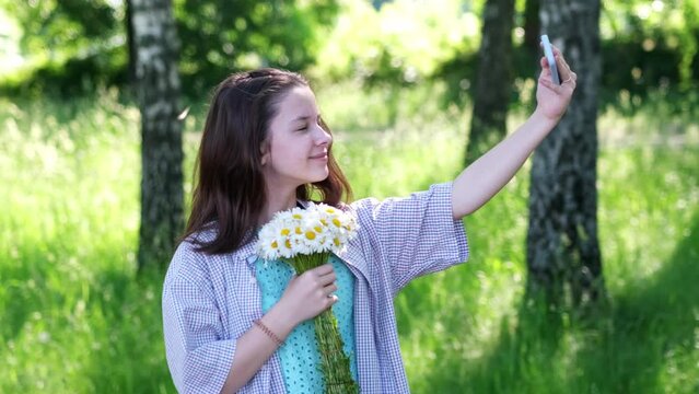 A young beautiful girl takes a selfie photo and video with her phone with a bouquet of beautiful daisies in her hand. Summer day and happy emotions on the face