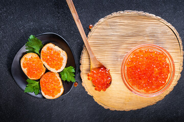 Log of wood on which lies spoon and glass bowl with red caviar next to it lies black plate with three sandwiches with caviar decorated with parsley. Red caviar on spoon and plates.