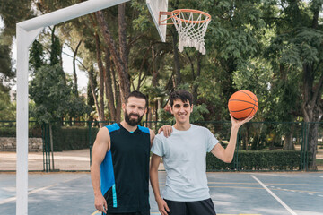Smiling young boys under a basket with a ball on a blue court. A couple of sporty men, one of them...