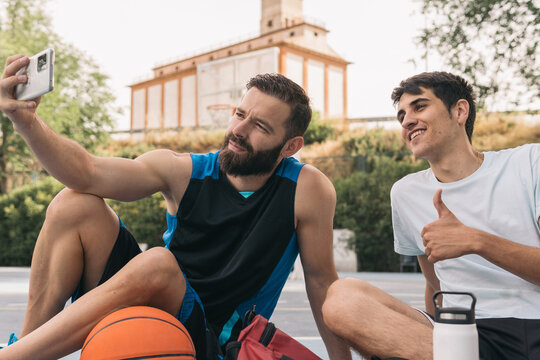 Smiling young guys sitting on a blue street basketball court and taking a selfie. A couple of sporty men, one of them holding a white phone and the other one with his thumb up taking a photo together.