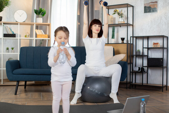 Young Sporty Mom Sitting On A Fitness Ball And Doing Sports Exercises With Dumbbells With Her Little Charming Girl In Sportswear Drinking Water From A Bottle While Standing After A Morning Workout.