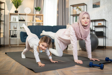 Body care, healthy lifestyle. Pretty muslim mother and her youth kid daughter doing stretching fitness exercise together in living room at home. Parent accompany children sport concept.