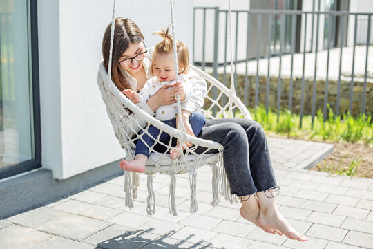 Happy mom and little baby girl are swinging on swing chair in backyard of house.