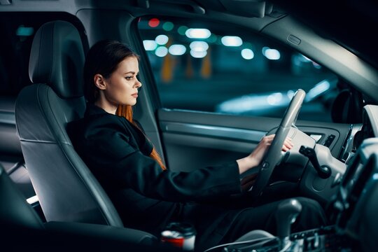 A Horizontal Photo From The Side, At Night, Of A Woman Sitting Behind The Wheel In A Black Shirt, Wearing A Seat Belt, Looking At The Road. Safe Driving Topics