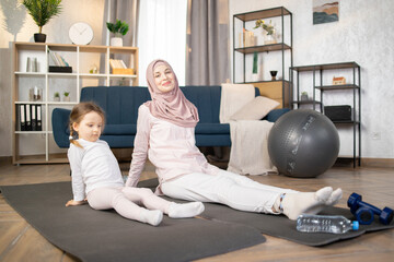 Young smiling muslim mother doing sports exercise at home, wearing hijab, together with funny...