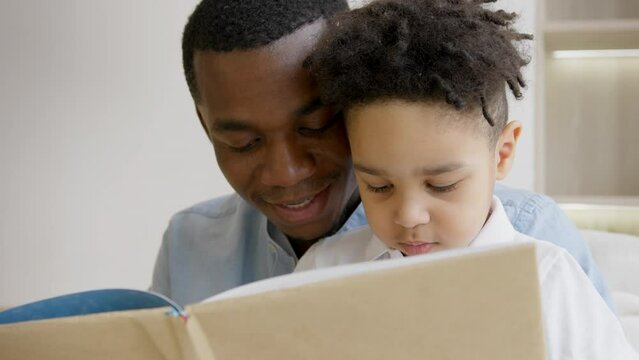 Single African American parent adult dad teach focused concentrated preschool child son reading book, learning education fairy tale story, bonding spend free time enjoy family lifestyle, home on sofa.