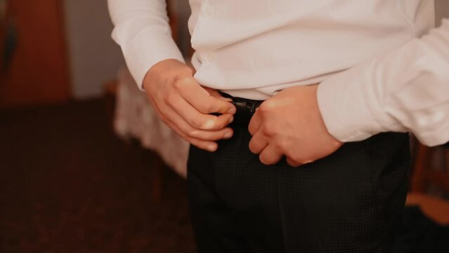 Khmelnytskyi, Ukraine - 08.08.2021: Stylish young man dressing in black suit fastening buttons white shirt jacket. man fastening belt in trousers. Close-up. Interior. Sunshine. Steady. Groom morning