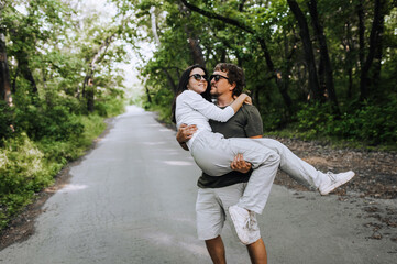 A bearded long-haired stylish man holds his beloved hippie brunette woman in sunglasses in his arms and circles her in nature in the park. Portrait, photo of newlyweds, concept.