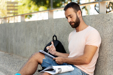 Handsome pensive man holding paper notepad, taking notes, writing resume sitting on the street....