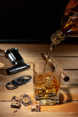 Pouring whisky from a bottle into a glass with ice cubes, cigar, cigar cutter and lighter