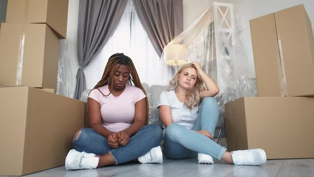 Sad relocation. Home moving out. Real estate. Two tired upset depressed diverse female friends sitting on floor in apartment with isolated furniture unpacked boxes.