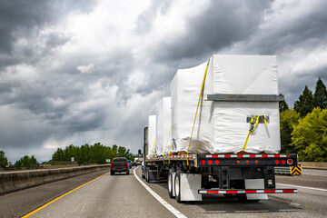 Powerful big rig semi truck tractor transporting heavy duty equipment on flatbed semi trailer driving on the multiline highway road with storm sky - obrazy, fototapety, plakaty