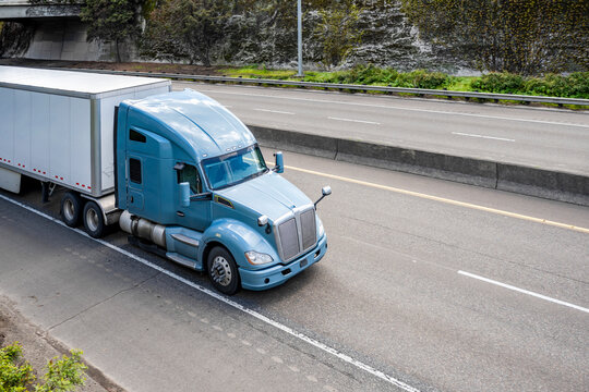 Powerful blue gray big rig semi truck transporting cargo in dry van semi trailer driving on the divided lower highway road