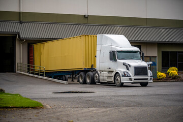 White big rig professional semi truck with container trailer unloading cargo standing in warehouse dock with gates