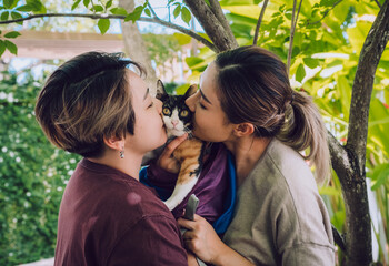 Happy lesbian couple with rainbow flag enjoy playing with their pet cat. Togetherness concept.